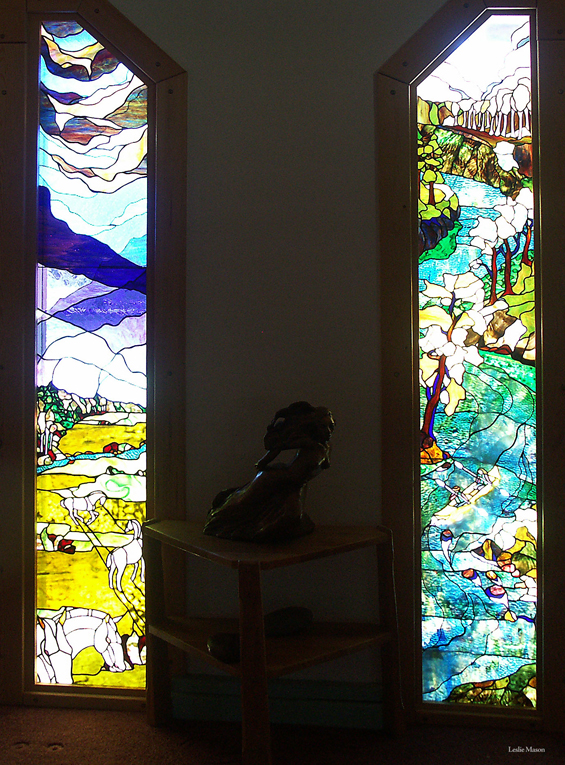 two tall windows, the left with horses grazing below a blue sky, the right with a river running through a forest