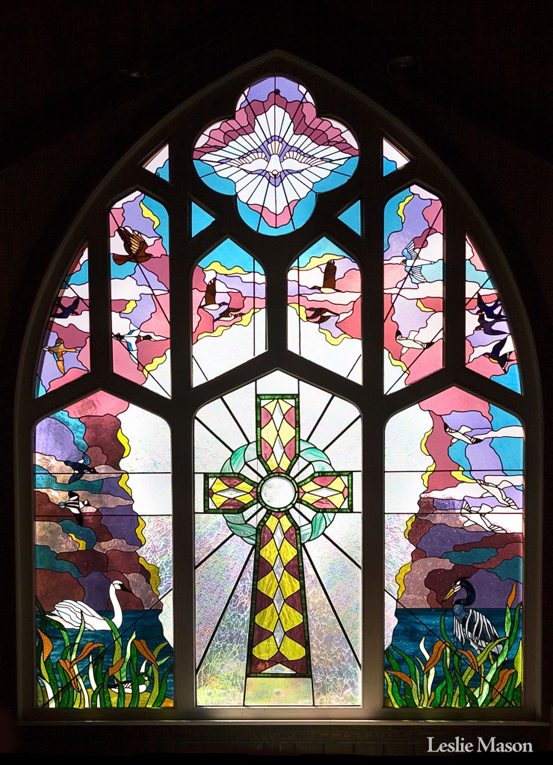 Large, multi-paneled church window with a cross at the center and birds flying and swimming in the surrounding panels